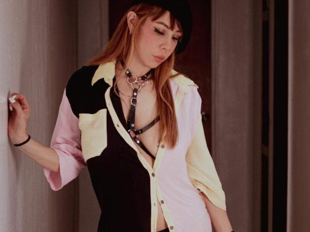 AriiannaHot with long red hair, wearing a black hat, pink, black, and yellow shirt, black harness, and metal collar, leaning against a wall, looking down.