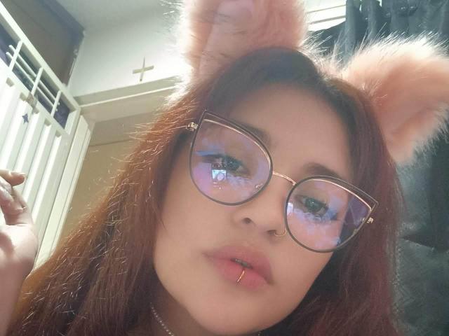 AgathaK with brown hair, cat ears, glasses reflecting a room, nose ring, and lip piercing, looking at the camera, light skin, subtle makeup, wearing a necklace.