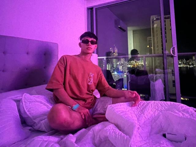 WillStarr, wearing a loose orange T-shirt and grey shorts, sits on a bed with white pillows, hands on knee, purple room lighting, city skyline reflected in glass door.