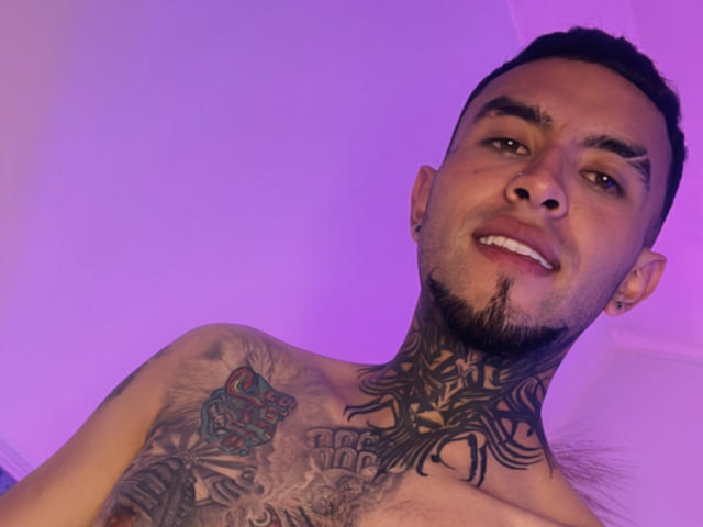 Shirtless LookBarber with dark tattoos on neck and chest, short black hair, goatee, smiling, purple lighting, ear studs, chest tattoo of dice, and intricate neck tattoo.