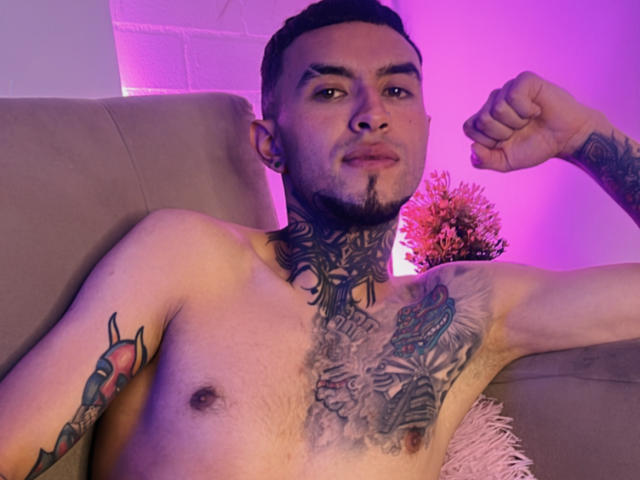 Shirtless LookBarber with tattoos, raised fist, and short dark hair, purple lighting, light skin, and a goatee, sitting on a beige couch, showing off chest and neck tattoos.