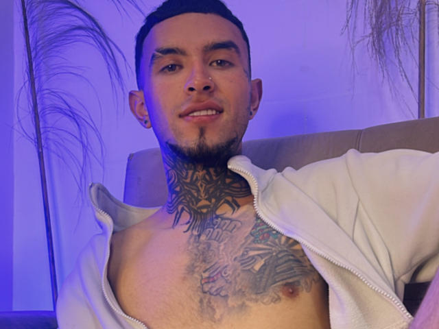 LookBarber with a goatee and tattoos, shirt unzipped to chest, revealing tattoos and a nipple, smiling, short hair, and a nose ring, under purple lighting.