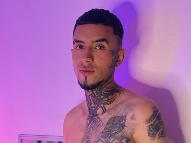 Tattooed LookBarber with short dark hair, beard, and earrings, shirtless, showing neck and chest tattoos, and a colorful chest piece, under purple and pink lighting.