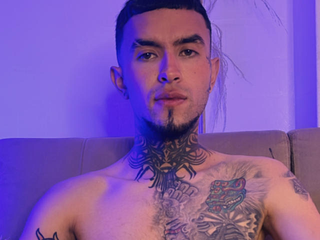 Shirtless LookBarber with dark hair, goatee, and tattoos on chest and neck, sitting under blue light. Visible chest tattoo of a red and green patterned design.