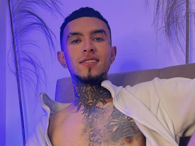LookBarber with tattoos on chest and neck, short black hair, beard, nose ring, white jacket, smiling, purple blue lighting, puffy chest, visible nipple.