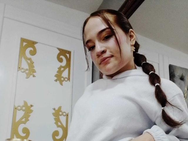 MadisonAngel with long brown braid, white sweater, gold earrings, and slight smile, showing subtle makeup, cute, and looking down with hands on hips.