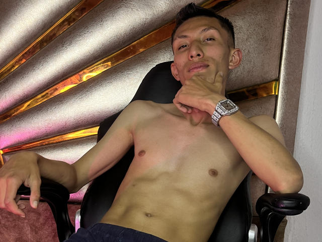 Shirtless BasthianFlitr with a slim, toned body, small ass, and a watch on his wrist, sitting in a chair, resting his chin on his hand, smiling.