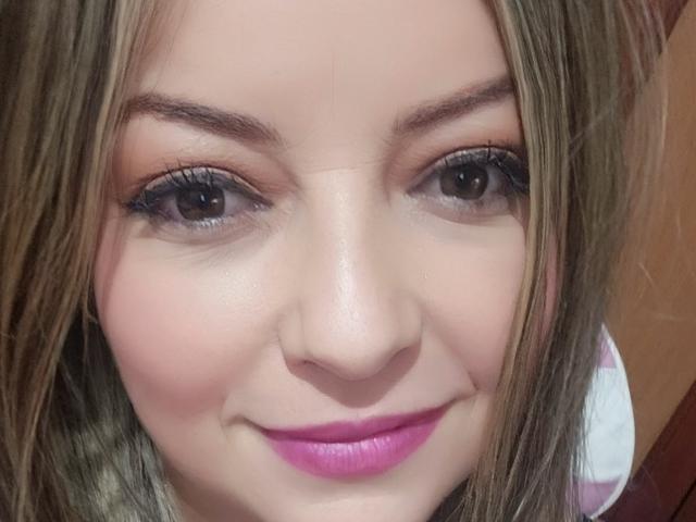 NatalyHami's face, with straight blonde hair, blue eyes, and pink lipstick, showing a subtle smile and lightly flushed skin. Boobs aren't visible, just her cute face.