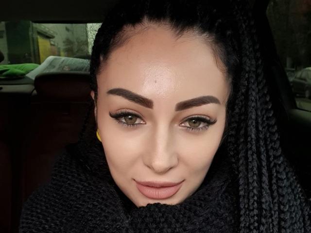 LilithLust has a shiny face with thick, dark eyebrows, green eyes, and full, pink lips. She's wearing black, braided hair and a black, textured scarf.