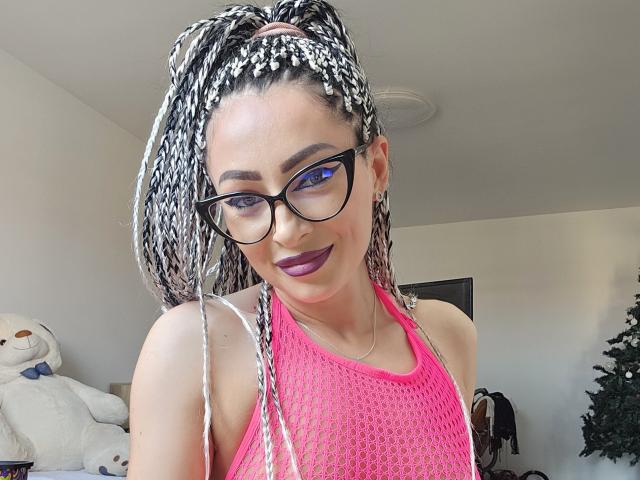 LilithLust with long braided white and black hair, purple lipstick, and black glasses, wearing a pink mesh top, smiling at the camera, showing her tits.