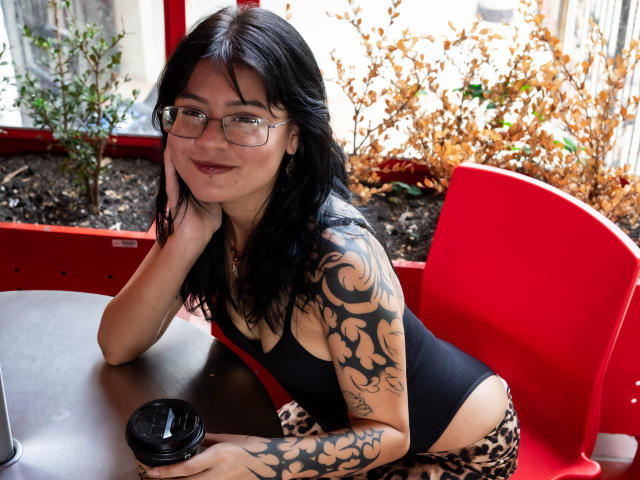 ElizabethRoux with black hair, glasses, and tattoos, leaning on a table, wearing a black tank top and leopard print pants, smiling, holding a coffee cup, seated in a red chair.