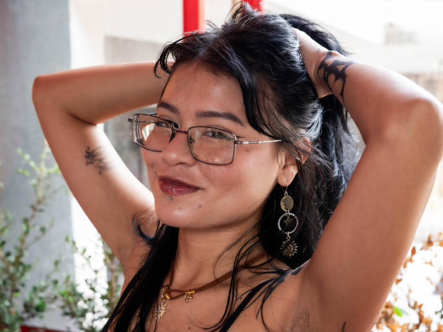 ElizabethRoux with glasses, black hair, arms raised, showing armpit tattoos, pierced lip, gold necklace, heart earrings, smiling, medium tits, Asian, sunlight.