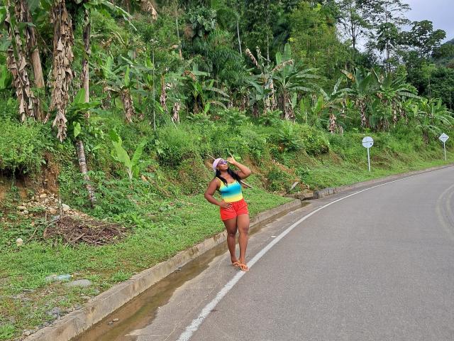 NattyN with a curvy ass, wearing a colorful tank top and red shorts, stands on a roadside, touching her head, with a lush green hill and road behind.