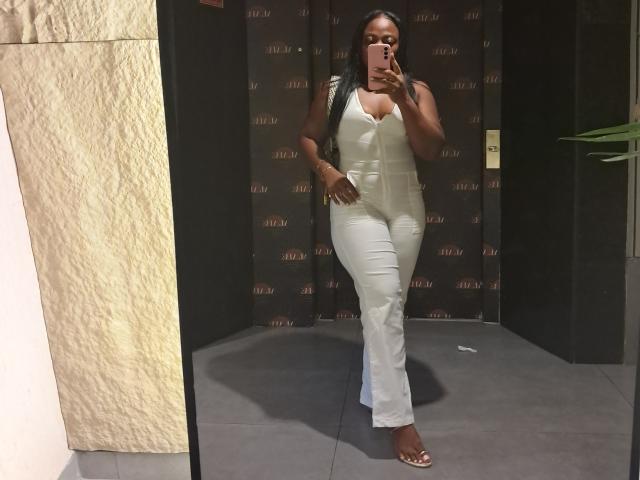 NattyN has dark skin, long black hair, and is wearing a white sleeveless bodysuit with white pants, taking a mirror selfie, showing her full ass and small tits.