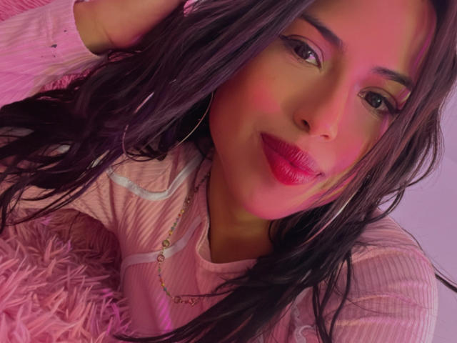 MyaaTyler with pink lipstick, long dark hair, hoop earrings, pink ribbed shirt, lying on pink fluffy pillow, smiling softly, pink and purple lighting on face.