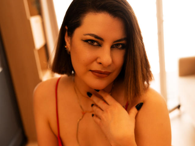 EmilyAmore with shoulder-length brown hair, red spaghetti strap top, black nail polish, and a subtle smile, touching her chest. Warm, soft lighting. Her tits are visible.
