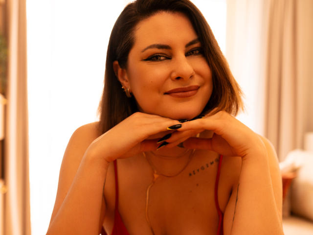 EmilyAmore with short brown hair, black nails, gold necklace, red top, and subtle smile, hands resting on chin, warm light, soft smile, cute, attractive.