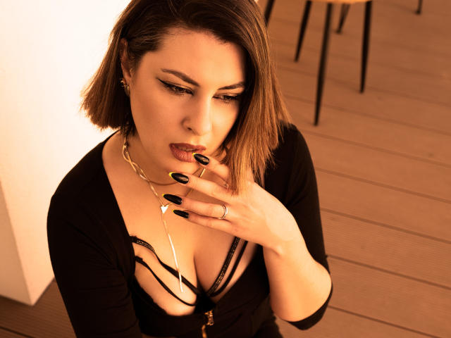 EmilyAmore met kort bruin haar, zwarte nagels en een diep decolleté, die op een gouden ring zuigt, gekleed in een zwarte jurk en een gouden ketting, en verleidelijk naar beneden kijkt..