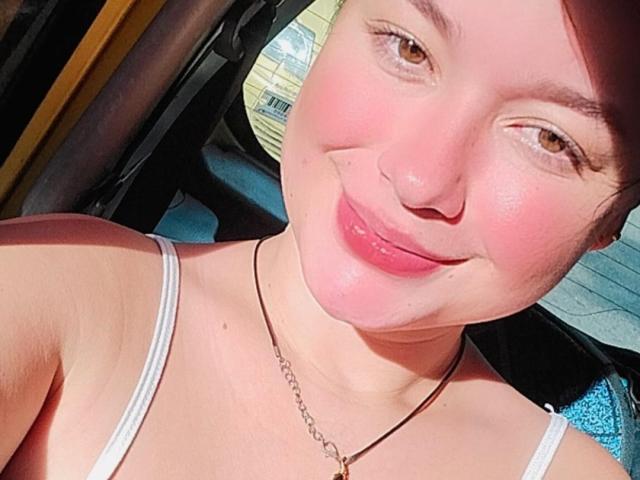 IsaLoverX, met roze wangen, bruine ogen en een neusring, lacht naar de camera, draagt een witte top met bandjes en een ketting, in een zonnige auto..