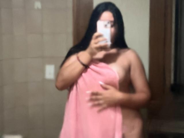 LyannaVega mit langen, schwarzen Haaren, nackt, bedeckt ihre Brüste mit einem rosa Handtuch und hält ein Handy in einem unscharfen Selfiemirror. Ihre Hand liegt auf ihrem Vulva..