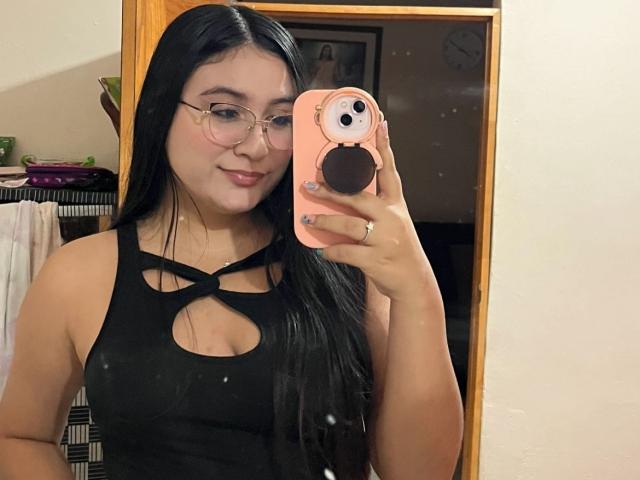 Lyanna Vega, con lunghi capelli neri, occhiali e una top nera con aperture, tiene in mano un telefono rosa con un adesivo carino sul retro e sta scattando un selfie allo specchio. Il suo seno è visibile..