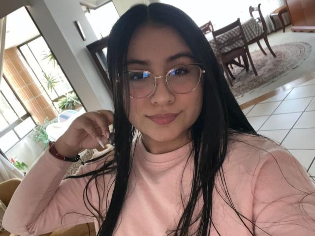 Lyanna Vega mit langen, schwarzen Haaren, Brille und einem pinken Pullover, lächelt mit leicht geschürzten Lippen und zeigt dabei ihren Hintern, wobei sie mit der Hand ihr Haar berührt. Innenaufnahme, helles, natürliches Licht..
