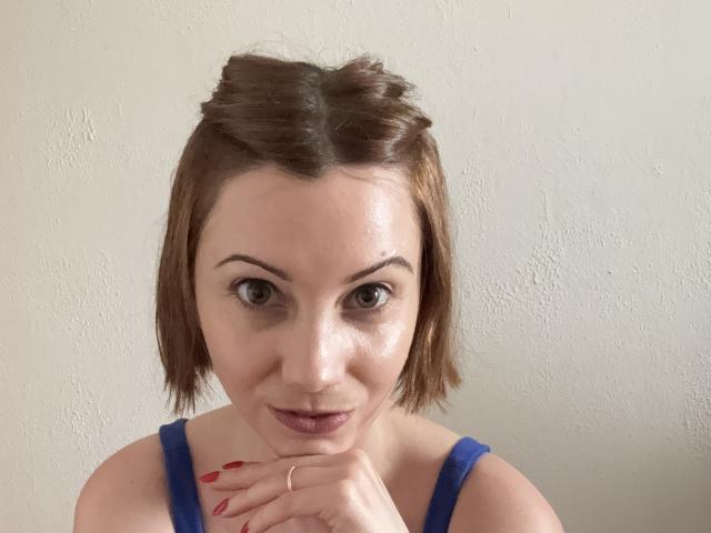 AfroditaMoore with short brown hair, braided top, blue top, red nails, ring on finger, looking at camera, slight smile, white wall in background.
