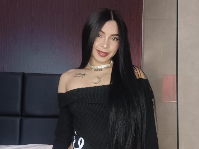 MistressAsshley with long black hair, off-shoulder black ribbed top, choker, and tattoo, smiling at camera, showing small tits and a crescent moon tattoo on her right chest.