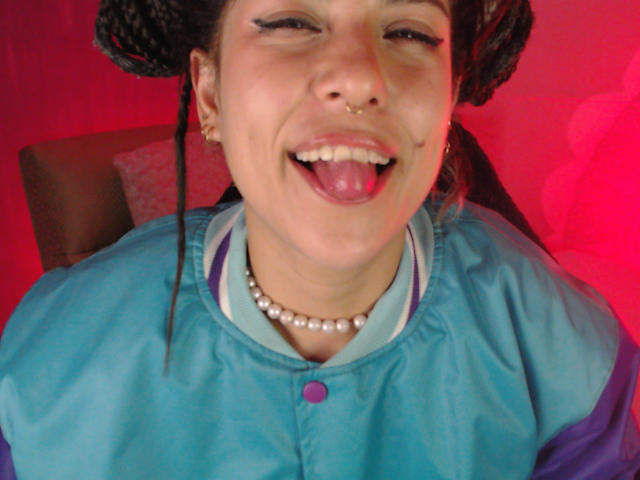 HellenK with braided hair, nose ring, and pearl necklace, tongue out, smiling, wearing a blue and purple jacket, small earrings, and a button-up shirt with a purple button.