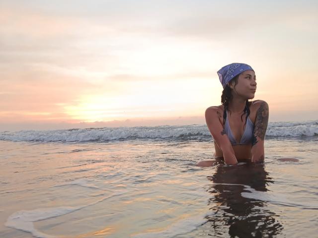 HellenK with wet brown hair, wearing a blue bikini and bandana, has a tattoo on her right arm, standing in shallow water at sunset, looking to the side.