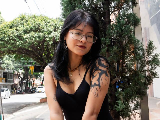 ElizabethRoux with black hair, glasses, and a black tank top, showing a tattooed arm, standing next to a tree, looking at the camera with a slight smile.