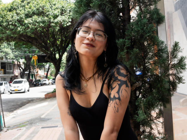 ElizabethRoux with black hair, glasses, and a tattooed arm, wearing a black tank top, standing by a tree, smiling with her tits slightly visible, wearing gold jewelry.