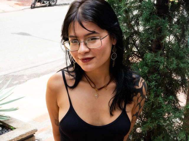ElizabethRoux with black hair, glasses, and a nose piercing, wearing a black tank top, showing tattoos on her right arm, and a necklace, standing by a green bush.