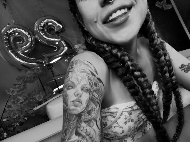 Schwarz-weißes Foto von HellenK mit geflochtenen Haaren, Nasenring, lächelnd, mit einem detaillierten Tattoo eines Frauenansichts auf ihrem Oberarm, die ein Oberteil mit Blumenmuster trägt..