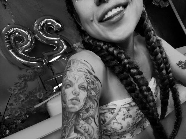 HellenK, avec des cheveux tressés, souriante, montrant un tatouage détaillé du visage d'une femme sur son bras droit, portant un haut fleuri, et des ballons métalliques en forme de "25" en arrière-plan..