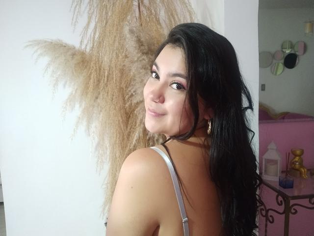 NicoletFoxy met lang zwart haar, gekleed in grijze lingerie met veters, die haar billen en rug laat zien, lachend over haar schouder, met pluizig beige gras op de achtergrond..