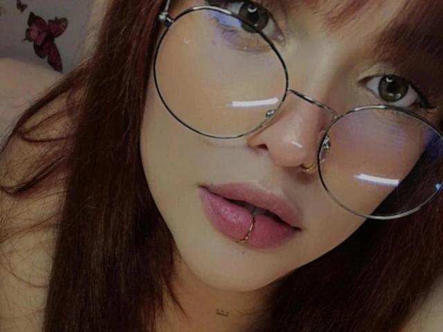 AgathaK with round glasses, red lipstick, and a lip ring, looking into the camera with her mouth slightly open, showing a small tattoo below her chin.