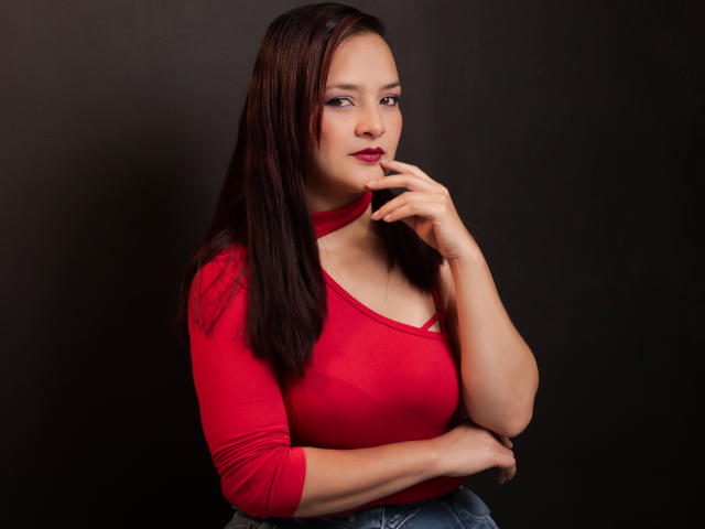 EvaLaura with dark brown hair, red shirt, and blue jeans, touching her chin with a finger, showing off her boobs and a confident smile. She's cute and sexy.