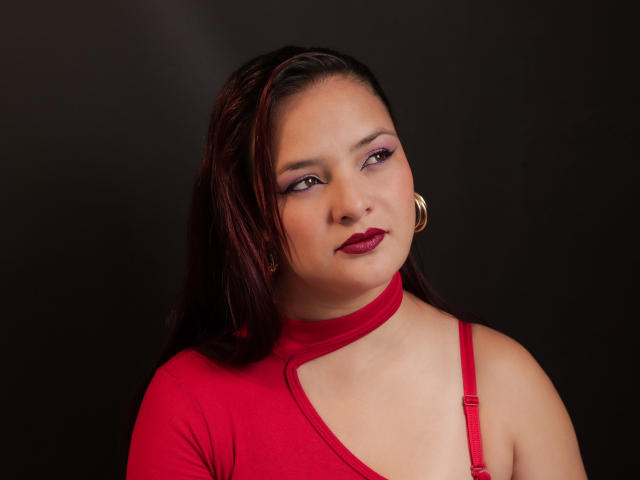 EvaLaura with dark brown hair, red lipstick, and red dress, showing her tits in a sleeveless top, gold hoop earrings, and a slight smile against a dark background.