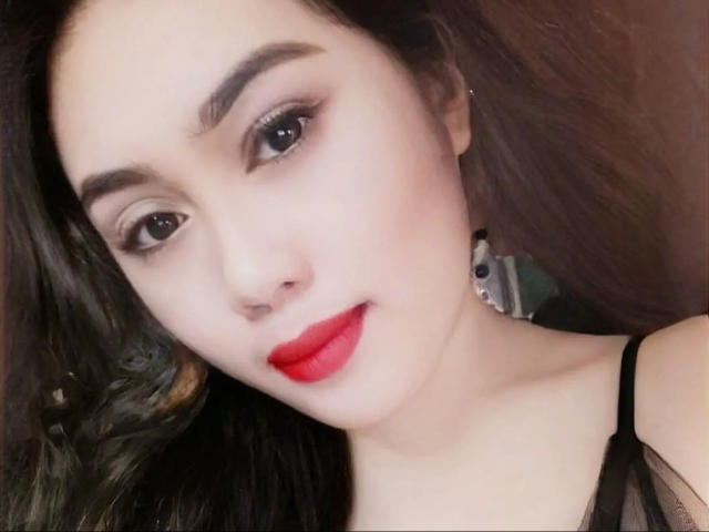 UrLadyXREM-hot with long black hair, wearing a white shirt, red tassel earrings, and a slight smile, touching her hair. She has light skin and red lipstick.