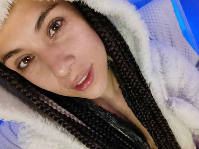 HellenK with long braids, nose ring, and choker, wearing a fluffy white hoodie, smiling, and showing teeth, has a cute face and is on a white textured blanket.