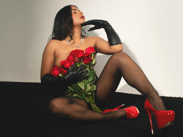 LindaFrancoo with black gloves, red high heels, black polka-dot tights, and red roses, holding them against her bare tits, sitting on a black surface, finger on chin.