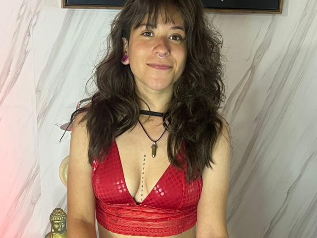 EveeDreams with wavy brown hair, nose ring, pink earrings, red lace bralette, black choker, necklace, smiling, medium tits, white marble wall, gold statue in background.
