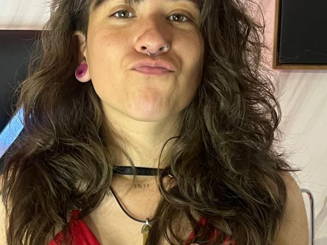 EveeDreams has wavy brown hair, a nose ring, pink ear gauges, and a slight pout. She's wearing a red top with a black choker. Her tits are small.