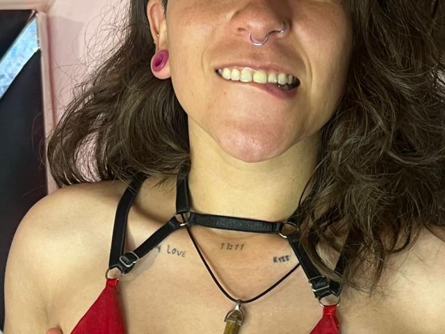 EveeDreams with wavy brown hair, nose ring, and pink ear gauges, smiling, showing teeth, wearing a black strappy top, red fabric, and black necklace, has "Love" and "Trust" tattoos.
