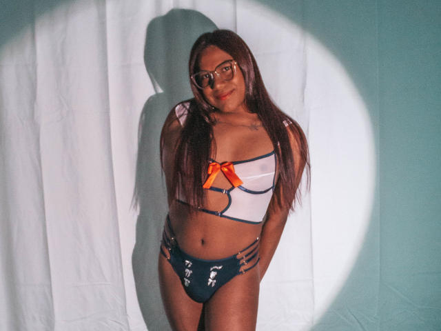 KimmySolange with long brown hair, glasses, and tan skin, wearing a white and orange lingerie top with cutouts, and blue panties with white fish designs, standing in shadow.
