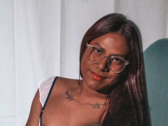 KimmySolange with dark brown hair, glasses, and a tattoo under her chin, smiling at the camera, showing her tits in a white top with a green strap.