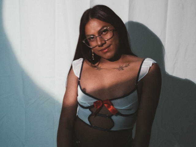 KimmySolange with dark hair, glasses, and a small red bow on her white top, showing her tits, standing in shadow, tattoos on her chest.