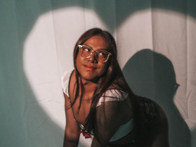 KimmySolange with long brown hair, glasses, white top, and necklace, leaning forward, shadow of her head on teal wall, smiling slightly, showing small tits and cute ass.
