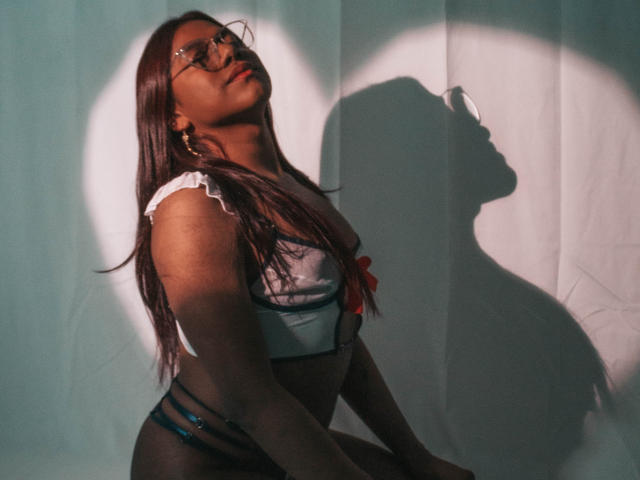 KimmySolange with dark brown skin, long red hair, glasses, white frilled top, red bow, black strappy bottoms, shadow on white curtain, sunlight highlighting her, looking up, confident.